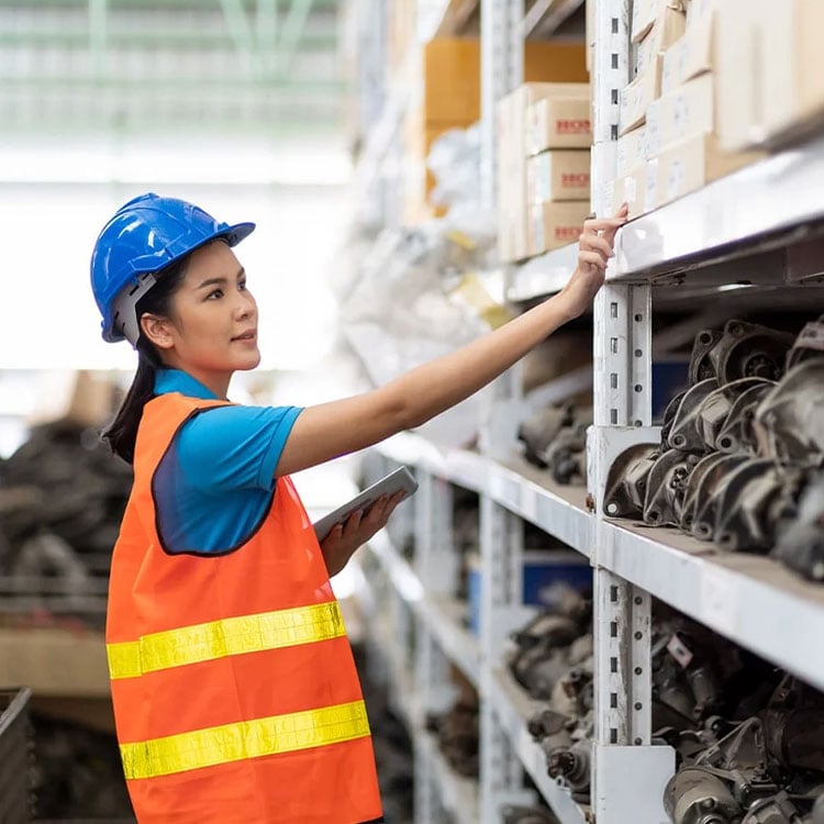 A woman in an orange vest and hard hat stands confidently in an Irish warehouse, looking for Automotive parts using KCS's automotive software