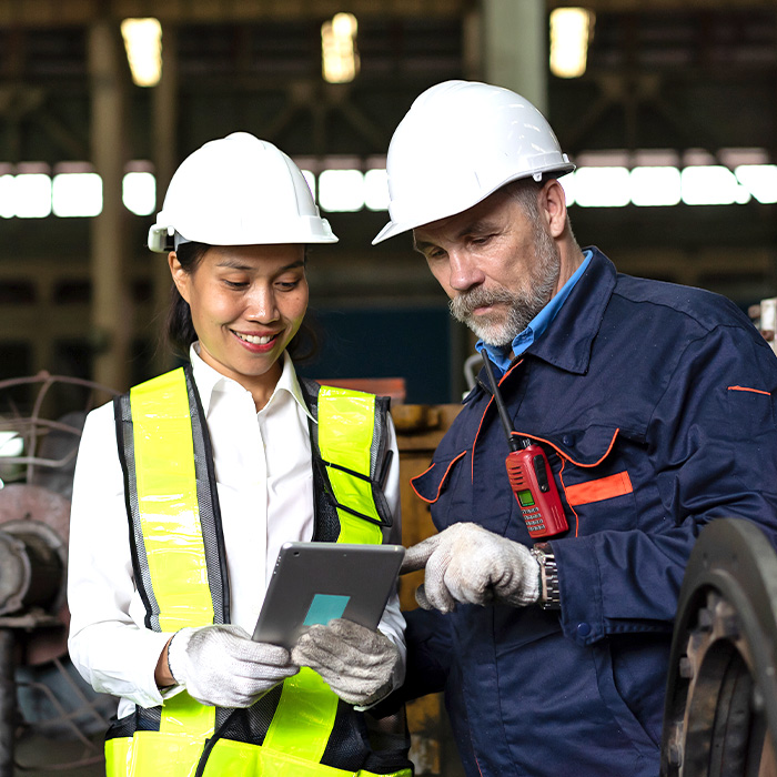 Two individuals in hard hats and safety vests using a tablet to manage automotive inventory.