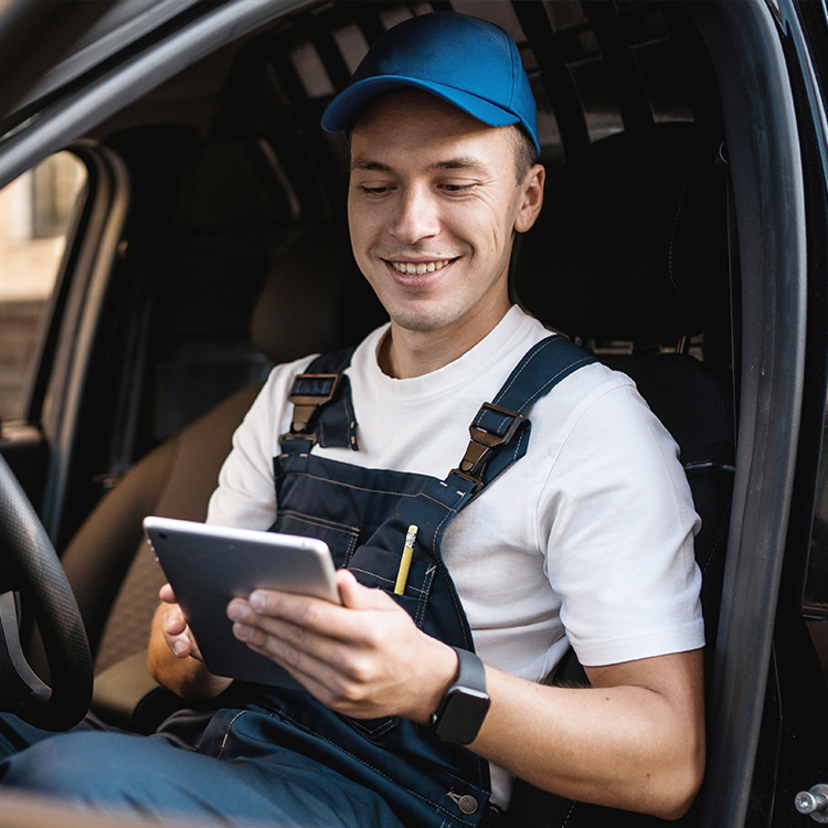 A man in overalls sits in the driver's seat of a car, holding a tablet. He appears focused and ready for a delivery