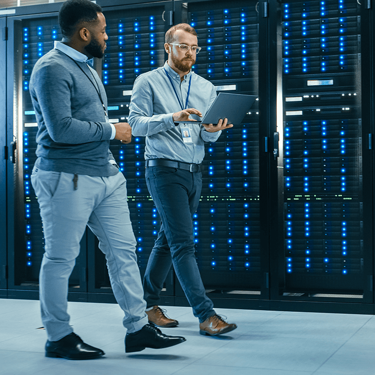 Two men walking in a server room looking at a laptop.