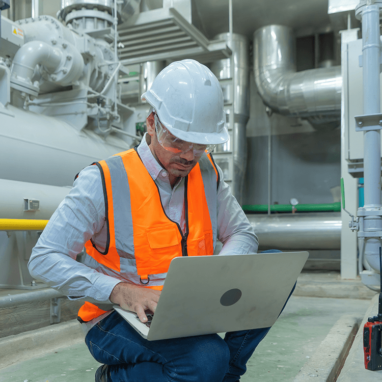 A man in an orange vest and hard hat sitting on a laptop, working in an Irish warehouse.