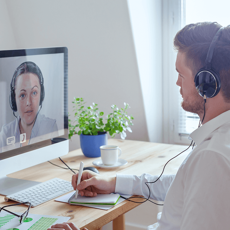 A man wearing headphones sits at a desk with a computer, engaged in a meeting with his colleague.