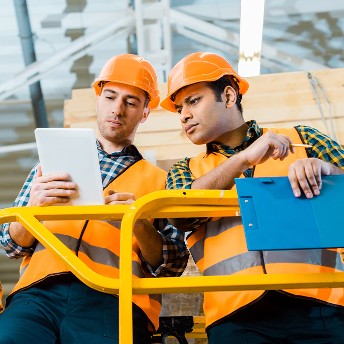 Two men in hard hats analysing data on a tablet to enhance efficiency in their merchant warehouse operations.