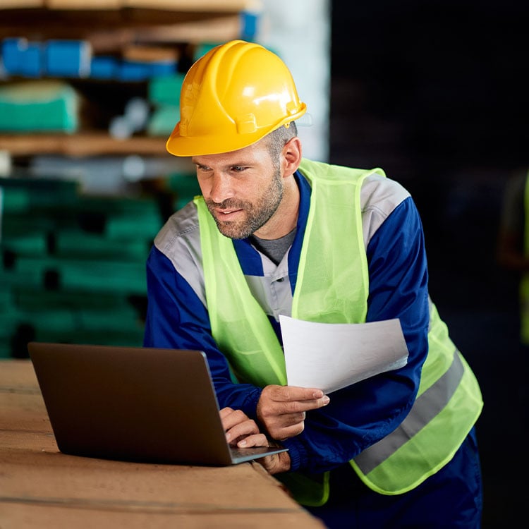 A worker in safety gear operates a laptop in an Irish warehouse, ensuring efficient timber warehouse management.