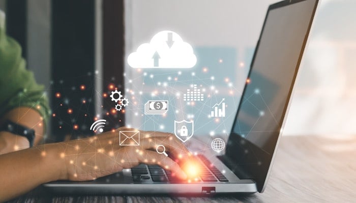 A person working on a laptop with a overlay of icons such as a cloud, wifi, email, shield and the internet icon signifying cloud computing.