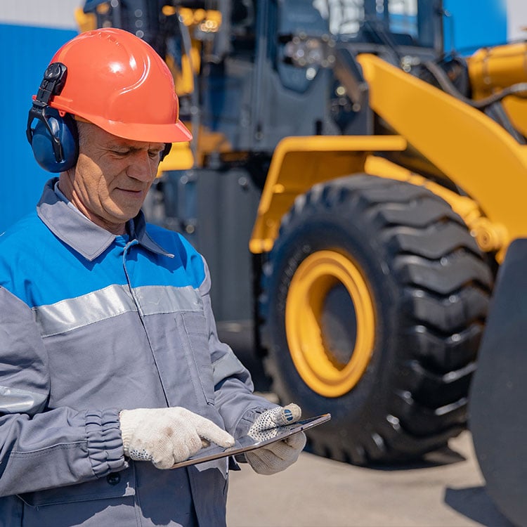 A person in a helmet holding a tablet, with a backhoe visible in the background.